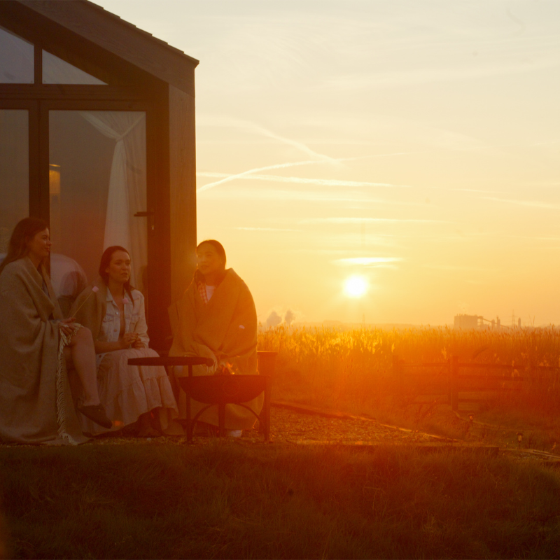 Three women roasting marshmallows over a fire pit at sunset