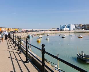 People walking along stone pier. Bright blue sky. Tide in with boats in the harbour. Gallery and residential buildings at the start of the pier
