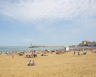 Looking across sandy beach with people sat or playing in the sea. Stone pier in background with flats, businesses and gallery building. Blue sky with some white cloud